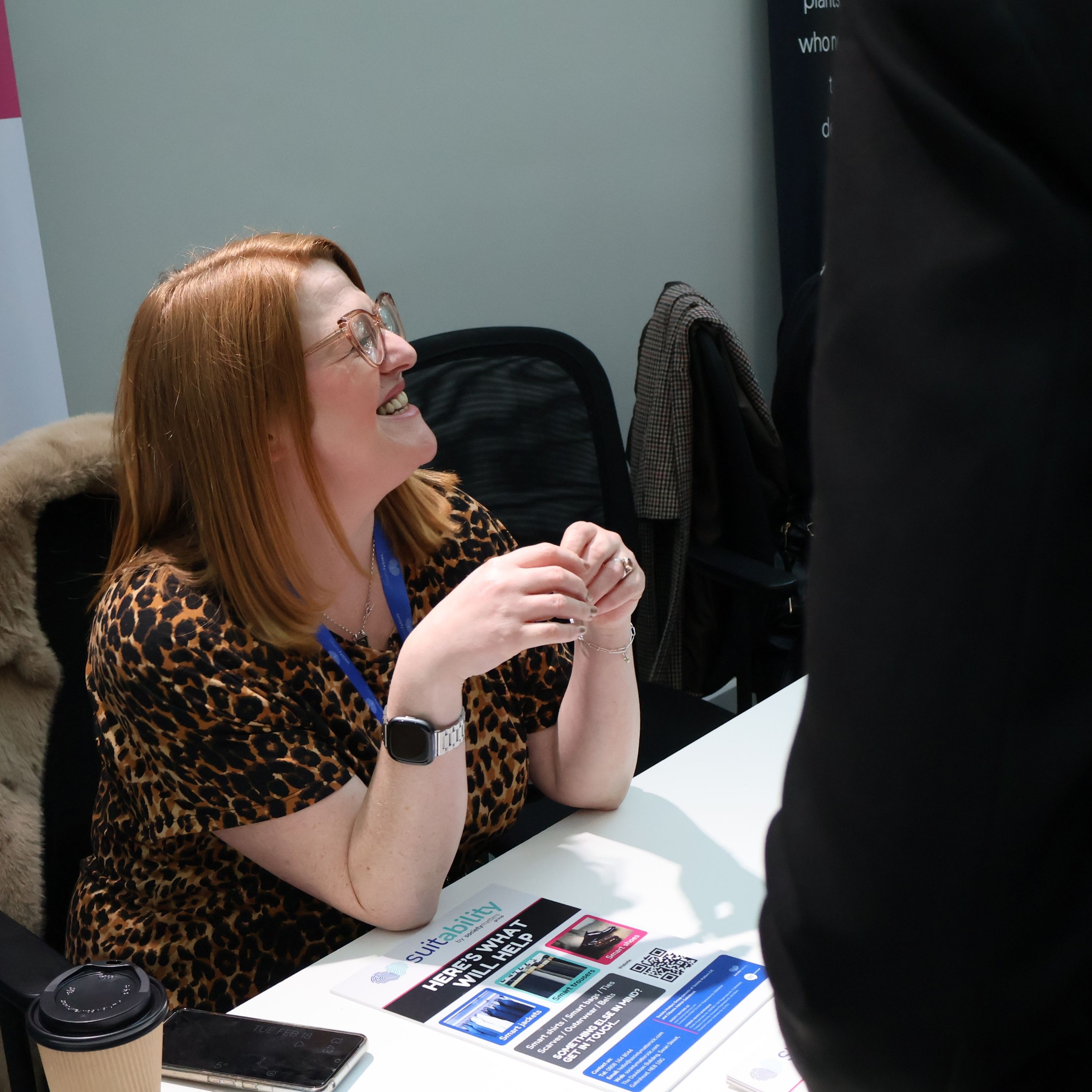 A woman with red hair, glasses, and a leopard print top sits at a table, smiling and engaging in conversation with a standing person. She is wearing a blue lanyard and a smartwatch. The table has flyers, a phone, and a coffee cup.