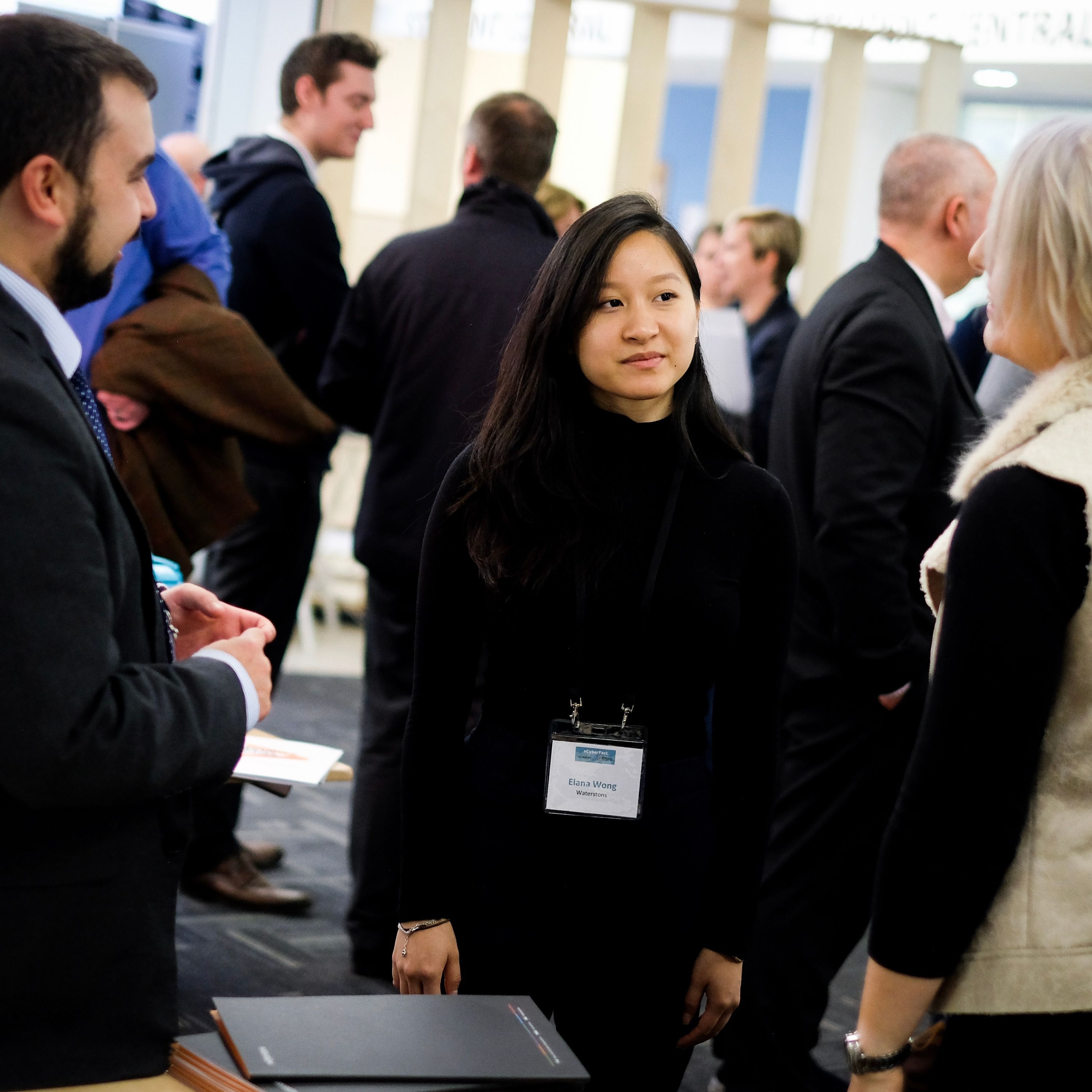 A professional networking event with people engaged in conversations. A woman in a black turtleneck and name badge stands in the center, listening to another attendee. Other people in business attire are in the background.