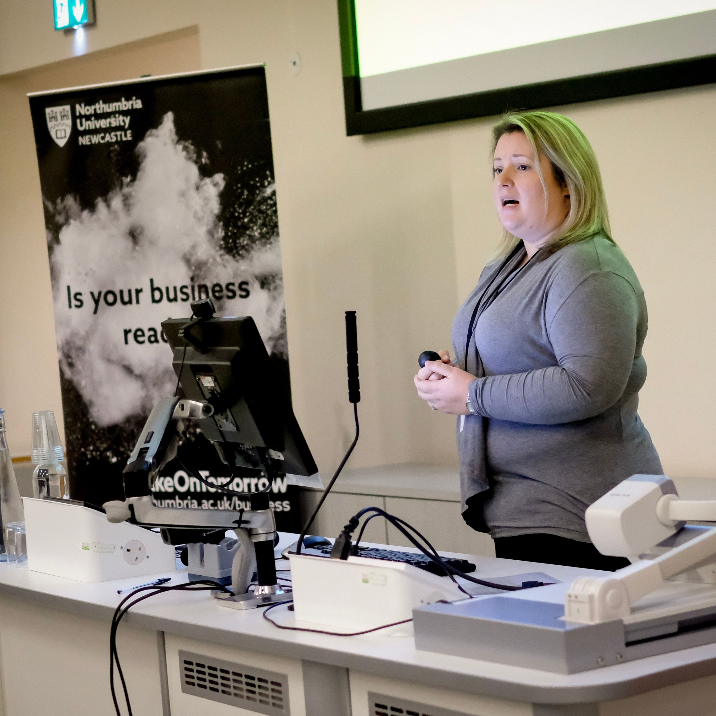 A woman with blonde hair wearing a gray sweater speaks in front of a table with a Northumbria University banner behind them. The table in front of her has a monitor, microphone and keyboard, all sitting on it.