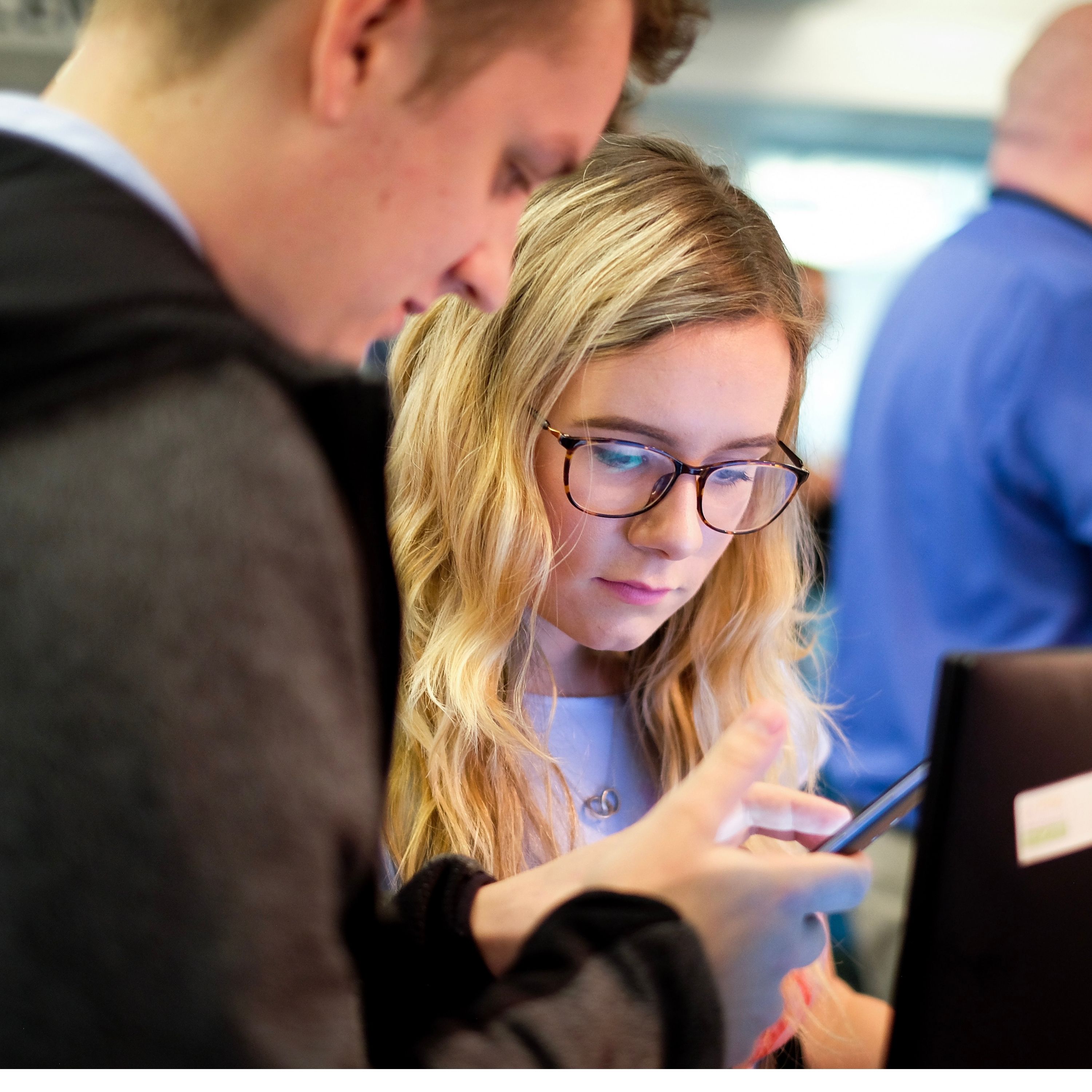 Close up shot of two people looking at a phone screen, a woman with blonde hair and glasses is in focus, while a man stands next to her holding a phone.