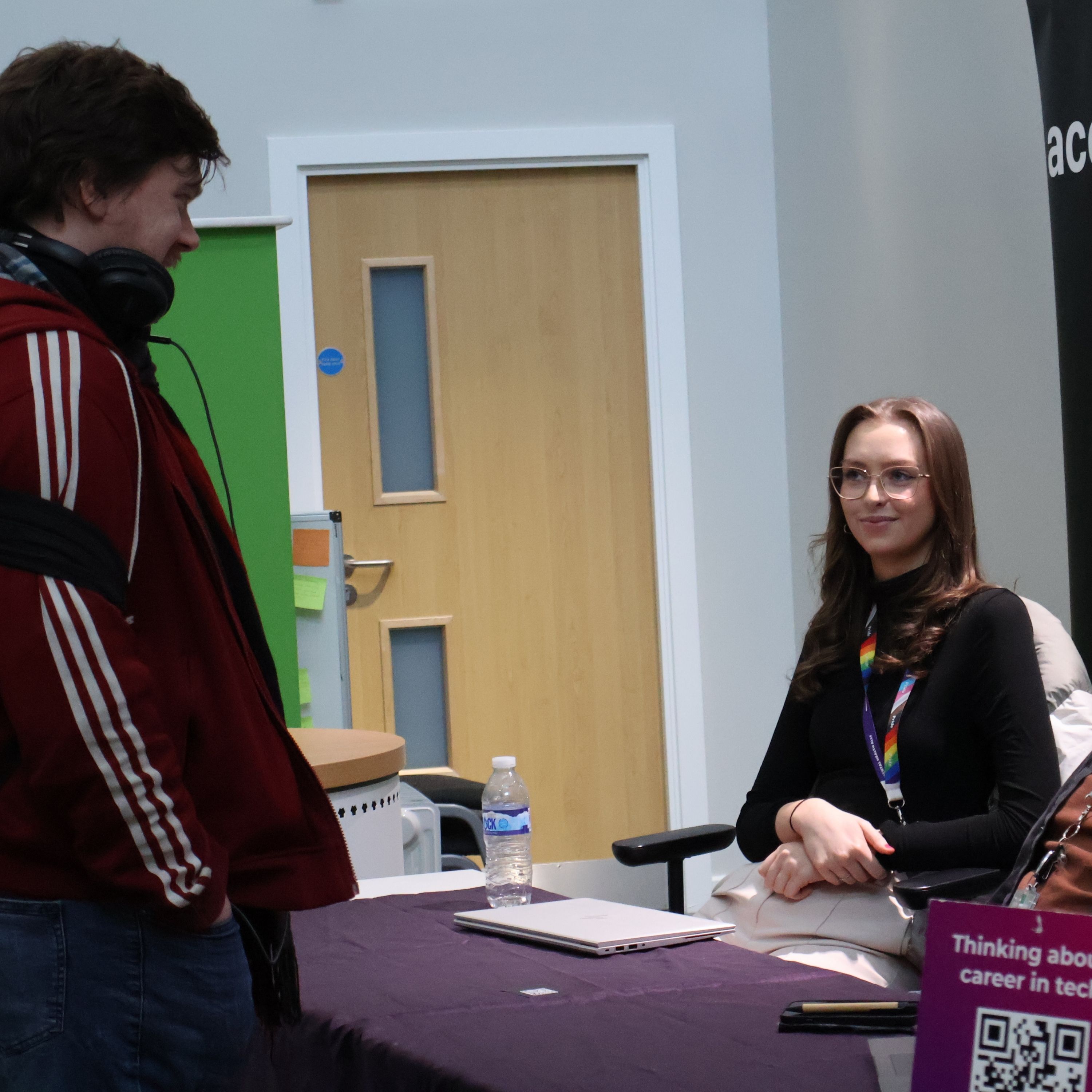 A woman wearing glasses and a black outfit sits at a table with a laptop and a water bottle, smiling while engaging in conversation with a man wearing a red jacket and headphones. They are at a career fair and a purple sign on the table mentions careers in tech.